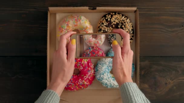 Las manos de las mujeres toman fotos con el teléfono inteligente de deliciosas rosquillas en la caja . — Vídeo de stock