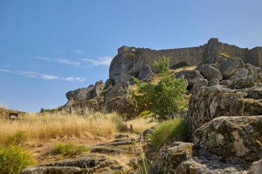Büyük kayalar ve açık mavi gökyüzünün altında vahşi bitki örtüsüyle çevrili antik taş kale duvarı..