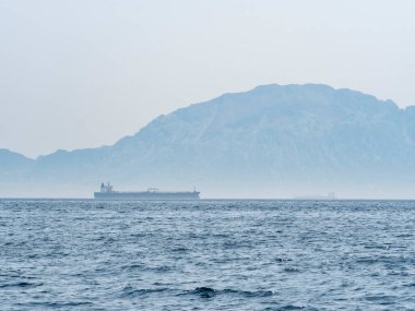 Büyük bir kargo gemisi Cebelitarık Boğazı 'ndan geçiyor. Arka planda Afrika kıyılarının sisli dağları görünüyor. Deniz ticareti ve küresel nakliye sahnesi