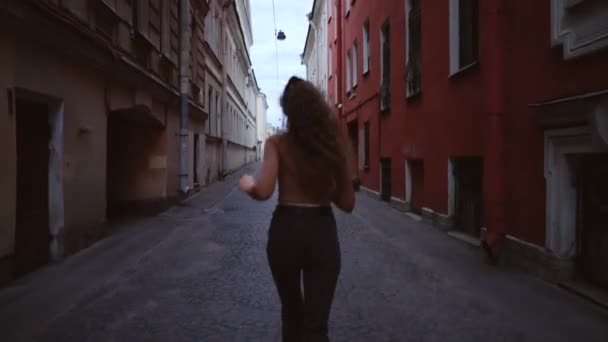 Junges schönes Mädchen dreht sich um und rennt auf eine europäische Straße. eine lächelnde fröhliche Frau blickt zurück. — Stockvideo