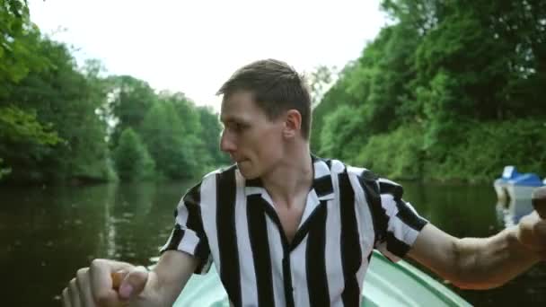 魅力的な若いブルネットの男は、ボートに座って、笑顔でオールで漕いでいます。緑の風景を背景にストライプのシャツを着た幸せな若者。夏のアウトドアアクティビティ. — ストック動画