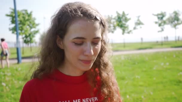 Curly menina atraente soprando em um dente de leão. Jovem mulher bonita fez um desejo. Senhora em uma camiseta vermelha em um fundo de grama verde. Uma bela flor feminina. Verão férias e feriados conceito . — Vídeo de Stock