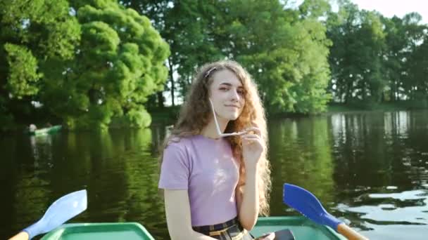 Jolie fille bouclée assise dans un bateau et souriante. Joyeux jeune femme dans un t-shirt violet tient des lunettes dans ses mains sur un fond de paysage vert. Activités de plein air estivales . — Video