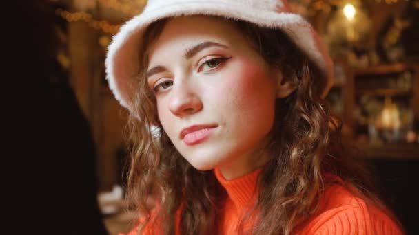 Attractive beautiful girl in a cafe looks cute. Young curly green-eyed woman in a stylish fluffy hat and orange sweater. Feminine Lady Close up — Stock Video