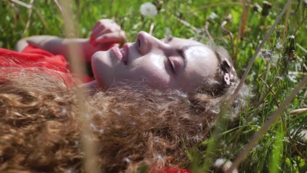 Кучерява приваблива дівчина лежить на зеленій траві. Молода красива жінка заплющила очі. Весела леді в червоній футболці. Концепція літніх канікул . — стокове відео