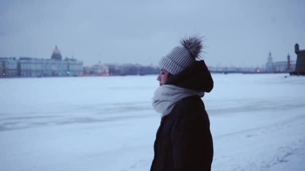 Menina nova em uma jaqueta quente e chapéu no frio. Jovem mulher em um contexto de snowdrifts e uma cidade nevada. Paisagem de inverno. Clima norte. Fundo bonito para letras ou texto . — Vídeo de Stock