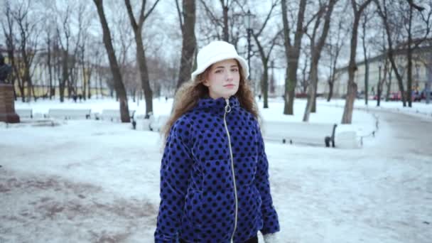 冬に魅力的なカーリーの女の子。白い雪を背景に青いジャケットと白い帽子をかぶった若い美しい女性。公園を散歩するというコンセプト. — ストック動画