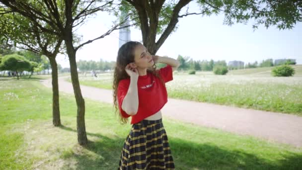 Curly menina atraente posando com as mãos. Jovem mulher bonita sorri e gosta com os olhos fechados sobre a natureza. Menina em uma camiseta vermelha e uma saia xadrez em um fundo de grama verde. Férias — Vídeo de Stock