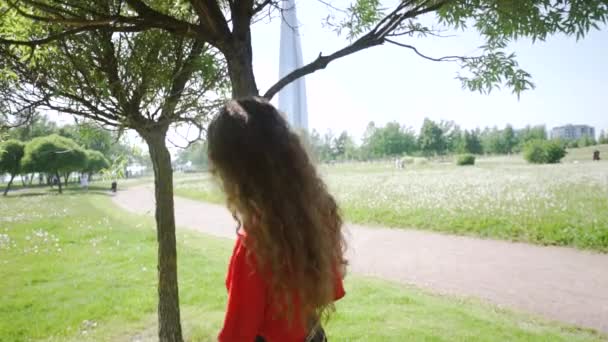 Curly attraktiv flicka spelas runt träden. Ung vacker kvinna virvlar och flirtar i naturen. Hona i en röd T-shirt och en rutig kjol på en bakgrund av grönt gräs. Sommarsemester koncept. — Stockvideo