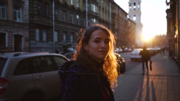 Lockiges attraktives Mädchen lächelt und sieht niedlich aus. junge hübsche Frau in einer europäischen Stadt im Hintergrund des Sonnenuntergangs. Mädchen in blauer Jacke aus nächster Nähe. das Konzept eines Abendspaziergangs. — Stockvideo