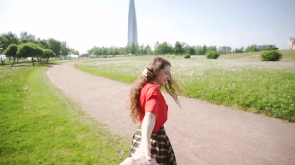 곱슬 매력적인 소녀는 남자의 손을 당깁니다. 젊은 예쁜 여자가 들판을 걷고 있습니다. 녹색 잔디의 배경에 빨간 티셔츠에 쾌활한 레이디. 컨셉은 나를 따르라. 여름 야외 활동. — 비디오