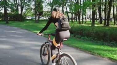 Güneşli bir günde parkta bisiklete binen beyaz, genç, kıvırcık kız. dış etkinlikler