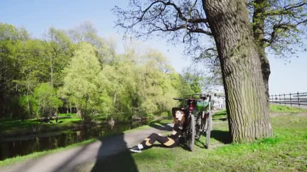 Joven hermosa chica sentada en el parque al lado del río con bicicletas. Emociones pensativas — Vídeos de Stock