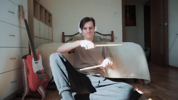 Young caucasian man with earphones sitting in his bedroom and playing in imagine drums with sticks 60 fps Slow motion — Stock Video