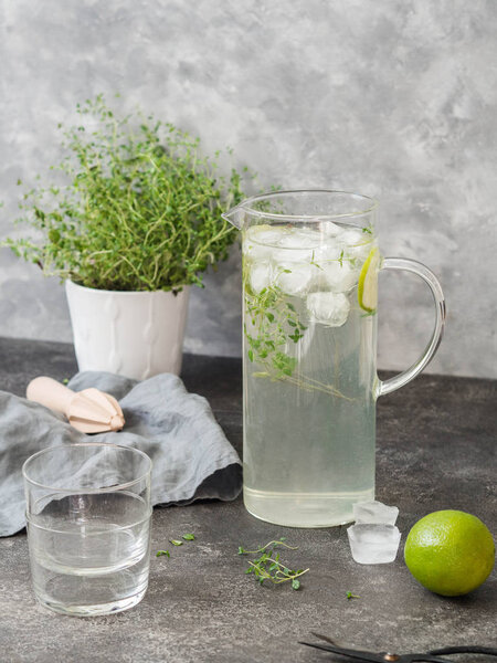Citrus lemonade with thyme in a glass jug, glasses and fresh thyme in a white pot on a gray table