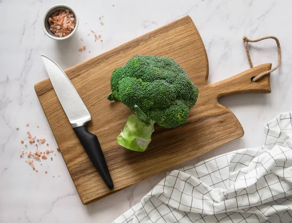 Broccoli on a wooden board, white towel and pink salt on a white marble table. Top view