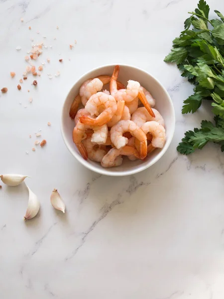 Shrimp in a white bowl, garlic, fresh parsley and pink salt on a white marble table. Top view. Healthy and diet dinner ingredients.