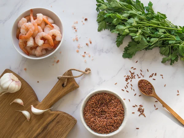Red shrimp in a white bowl, brown rice in bowl and in a wood spoon, garlic on wood board and fresh parsley on a white marble table. Top view. Healthy and diet dinner ingredients.