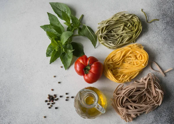 Different ingredients of Italian cuisine on a gray background. Multicolored paste or fetuccini, fresh green basil, red tomato, olive oil and spices on a gray background. Top  view