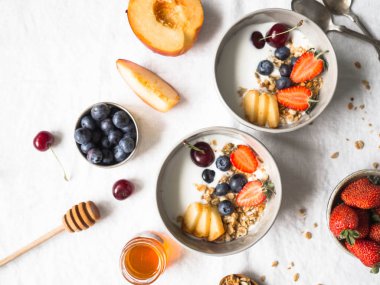 Ev yapımı granola bir kase yoğurt, şeftali, yaban mersini, kiraz ve çilek beyaz masa örtüsü ile bir tabloda. Beyaz bir arka plan üzerinde sağlıklı kahvaltı. Üstten görünüm. Kopya alanı