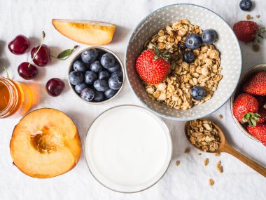 Ev granola bir kase, yoğurt bir cam, meyve ve çilek beyaz masa örtüsü ile bir tabloda. Sağlıklı kahvaltı için malzemeler. üstten görünüm.