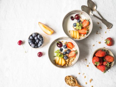 Ev yapımı granola bir kase yoğurt, şeftali, yaban mersini, kiraz ve çilek beyaz masa örtüsü ile bir tabloda. Beyaz bir arka plan üzerinde sağlıklı kahvaltı. Üstten görünüm. Kopya alanı