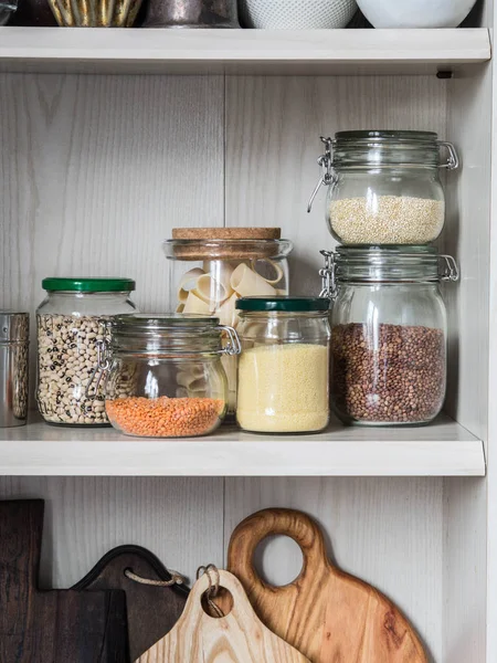 Shelf in the kitchen with various jars of cereals and kitchen chopping boards. Glass jars with pasta, lentils, couscous, beans and quinoa.