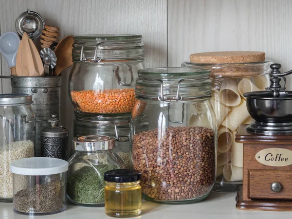 Shelf in the kitchen with various jars of cereals and kitchen tools. Glass jars with pasta, lentils, couscous, beans and quinoa. Coffee grinder, honey, spices, herbs, sesame