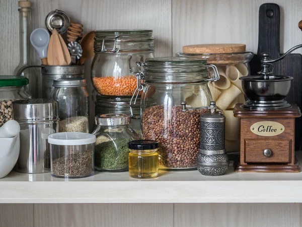 Shelf in the kitchen with various jars of cereals and kitchen tools. Glass jars with pasta, lentils, couscous, beans and quinoa. Coffee grinder, honey, spices, herbs, sesame