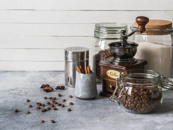 Divers ingrédients pour faire du café, bocaux en verre avec grains de café, sucre, moulin à café et laitier avec cannelle et étoiles d'anis sur fond gris. Espace de copie — Photo