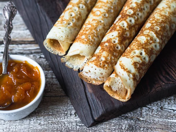 Russian homemade yeast pancakes rolled into a tube on wood board, apricot jam in bowl on wooden table. Traditional wheat pancakes for Shrovetide.