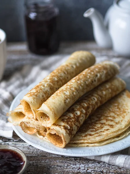 Russian homemade yeast pancakes rolled into a tube on white plate, jam and tea in the mug on the table. Traditional wheat pancakes for Shrovetide.