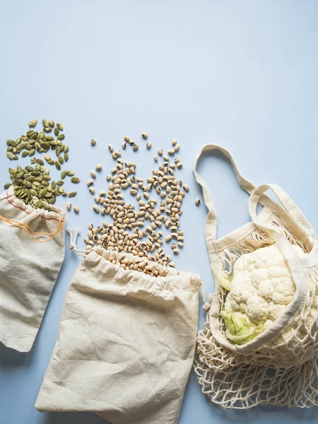 Eco-friendly beige shopping bags with cauliflower, beans, pumpkin seeds on a blue background. String bag with vegetables. Zero waste, no plastic concept.