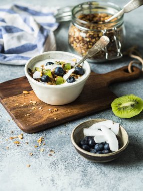 Ev yapımı granola yoğurt, yaban mersini, kivi, beyaz bir kase içinde Hindistan cevizi ile. Gri masada lezzetli Sağlıklı Kahvaltı