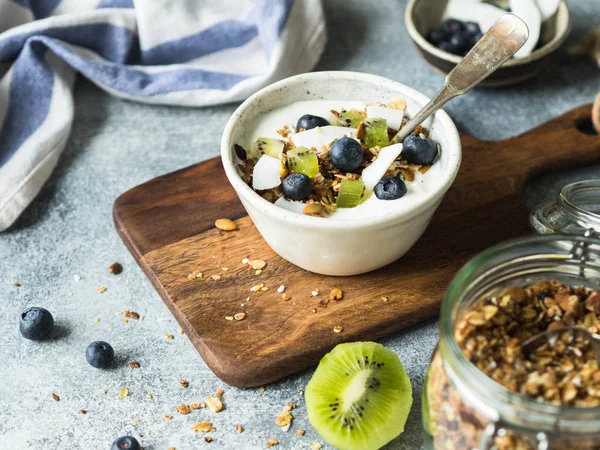 Granola casera con yogur, arándanos, kiwi, coco en un tazón blanco. Delicioso desayuno saludable en la mesa gris —  Fotos de Stock