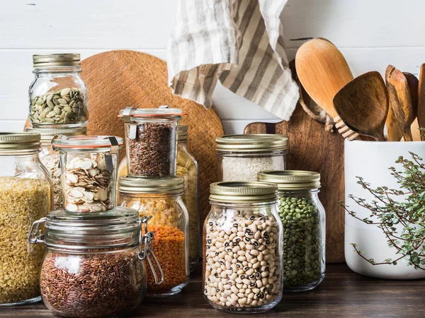 Vários cereais e sementes - ervilhas divididas, sementes de abóbora, feijão, arroz, macarrão, aveia, cuscuz, linho, lentilhas, fatias de amêndoa, bulgur em frascos de vidro na mesa na cozinha — Fotografia de Stock