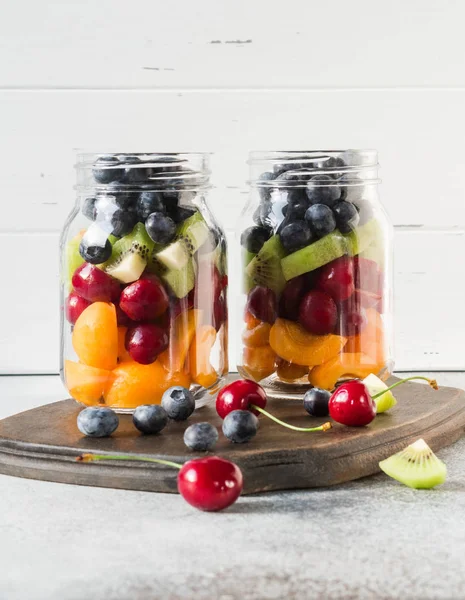 Two glass jars with berries and fruits. Fruit salad with apricots, kiwi, cherries and blueberries in glass jars on white background. Summer seasonal tasty vegetarian salad for a snack or lunch — Stock Photo, Image