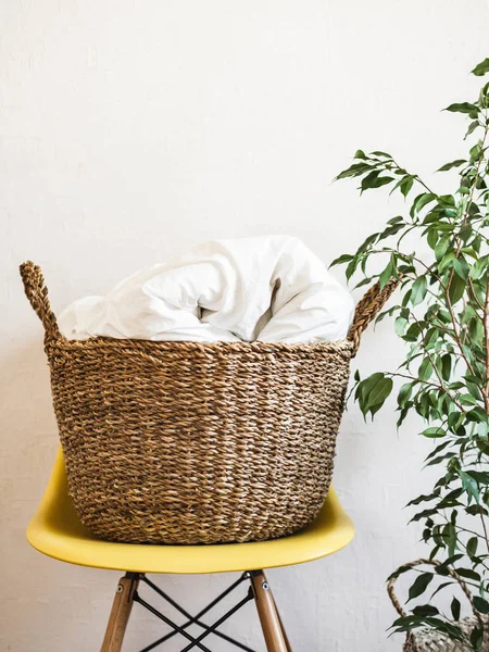 Grand panier en osier avec une couverture blanche sur une chaise jaune et une plante d'intérieur contre un mur blanc. Espace de copie — Photo