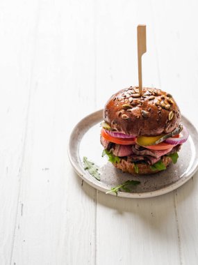 Biftek dilimleri, roka, domates, salatalık turşusu ve kırmızı soğanlı sandviç. Hamburger, çeşitli tohum ve beyaz ahşap arka planda seramik tabakta biftek.