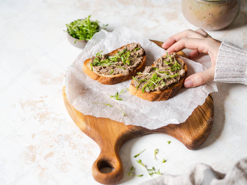 Two toasts chicken rillettes (pate) on white bread with sprouts on a wood cutting board and female hand takes toast on marble background. Copy space