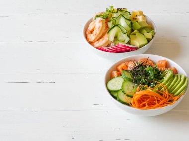 Beyaz ahşap arka planda deniz ürünleri, taze sebzeler, meyveler ve pirinçle iki farklı Hawaii poke kasesi.