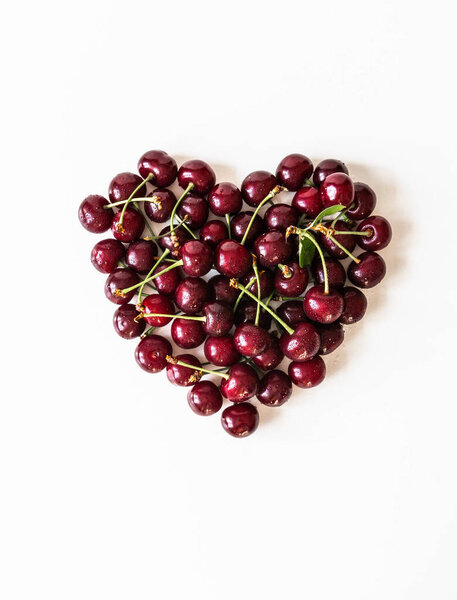 Red heart made from large ripe cherry berries with leaves and drops of water on white background. Top view