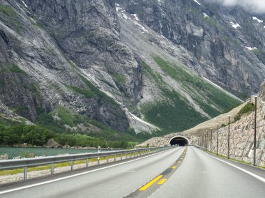 Çift sarı çizgileri olan boş bir asfalt yol ve göl kenarındaki dağda bir tünel. Bulutlu havada Norveç dağ yolu