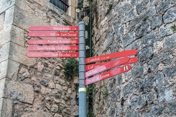 Ortaçağ Girona 'sında Banys Arapları, Katedral, Sant Feliu ve Arkeolojik Yürüyüş gibi tarihi mekanların yönlerini gösteren bir turist tabelası. Kırmızı oklar antik bölgenin taş duvarlarına monte edilmişti. Girona, İspanya - 28 Kasım 2021