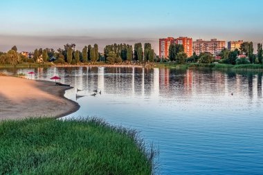 Ukrayna 'nın Poltava bölgesindeki Myrhorod tatil beldesindeki Khorol Nehri sahilinin akşam manzarası, sakin su yansımaları, kumlu sahil ve Sovyet döneminden kalma apartman arkaplanı