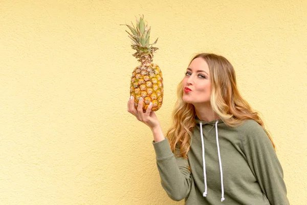 Sensual young blond woman with red lips leaning over to kiss a ripe pineapple she is holding in her hand over a yellow studio background with copy space