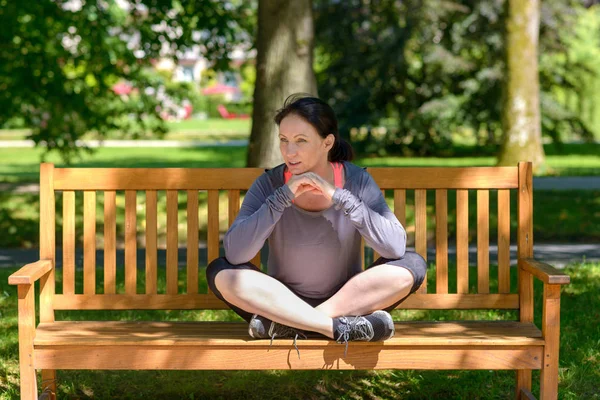 クロス脚側に彼女が見て喜んで笑っている緑豊かな緑の木々 の下で木造公園のベンチに座っている魅力的な運動しなやかな女性 — ストック写真