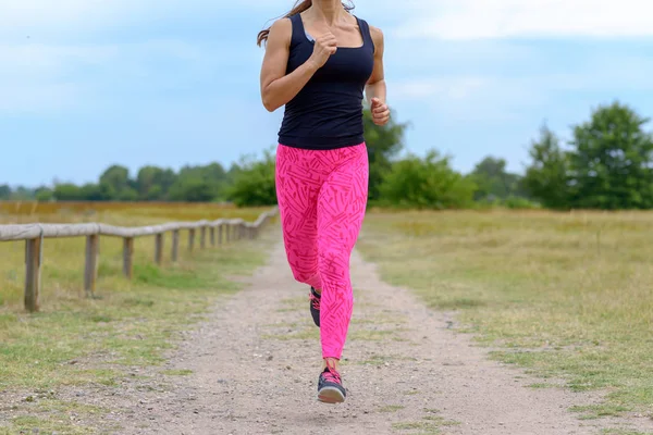Unidentifiable female runner with pink pants jogging along empty gravel road next to open green field