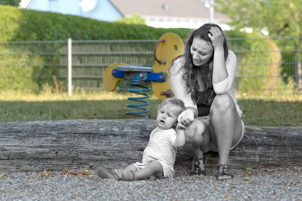 Troubled stressed young mother with her crying unhappy baby son outdoors in a playground in a conceptual image with de-saturated foreground