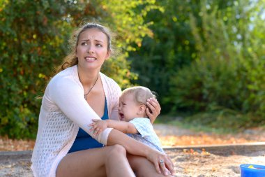 Stressed young mother comforting her crying baby son looking away with an anguished expression as they sit together in a playground outdoors clipart